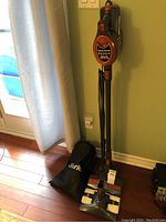 Full view of the Shark Rocket vacuum cleaner standing upright on hardwood floor next to a light green wall and a window with curtain. Includes vacuum unit and black fabric Shark dust bag on floor.