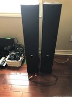 Front view of both black tower speakers on hardwood floor alongside cables