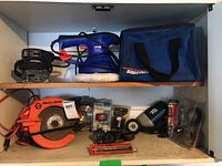 Full view of shelf showing Black & Decker circular saw, Craftsman palm sander, Black & Decker drill, Simoniz orbit polisher with blue carrying bag, and various accessories.