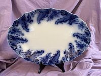 Full view of the antique glaze blue and white ceramic platter with scalloped edges on a black stand.