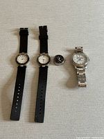 Four watches laid out: two identical black strap quartz wristwatches, one small black pocket watch, one silver linked metal wristwatch.