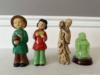 Four Chinese ceramic figurines displayed in a row: two children, an elderly man, and a green jade-like figure on wooden base.