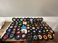 Top-down view of approximately 56 seven inch vinyl records spread out showing a variety of label colors and designs.
