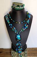 Front view of a display bust showing two beaded necklaces in blue and green tones with a large blue glass pendant and multiple wrap bracelets with mixed beads.