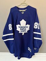 Front view of Toronto Maple Leafs blue hockey jersey with number 81 on sleeves and large white maple leaf logo with text 'Toronto Maple Leafs' on chest, hanging on hanger against wall.