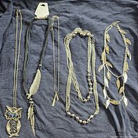 Five assorted silver tone necklaces laid out on a dark fabric background showcasing the owl pendant necklace, black cord feather charm necklace, geometric pendant necklace, triple-strand chain necklace with beads, and leaf charm necklace.