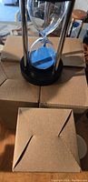 Five black frame sand timers with blue sand, each placed on top of or near individual cardboard boxes.