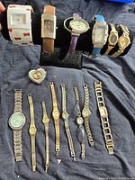 Full overview of 15 assorted watches with different strap materials and colors displayed on black cushion and cloth. Includes two cuff bracelets and heart-shaped desk clock.
