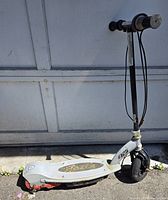 Side view of white Razor electric scooter leaning against a garage door showing the full scooter profile with handlebar, deck, and wheels.