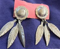 Pair of silver-toned feather earrings with a floral embossed round stud base and three feathers hanging from each earring.