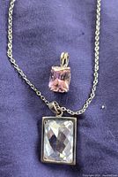 Close-up photo showing the two pendants, one clear rectangular faceted stone set in a silver frame, and one smaller pink faceted stone pendant on a silver-tone chain