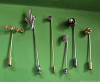 Six decorative stick pins laid out on green background, showing all pins and their varied designs