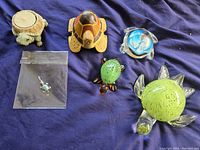 Six turtle items displayed on dark fabric: three glass turtles, one wooden turtle, one resin tea light holder, and one turtle pendant in bag.
