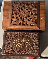 Top view of two rectangular carved wooden boxes showing the detailed floral carvings on the lids. One box has an oval floral medallion with gold accents, the other a square floral lattice design.