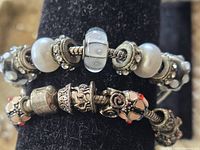 Close-up showing two silver-tone charm bracelets; the first with white and black beads, some with rhinestones, the second with detailed floral and ornamental charms including red enamel accents.