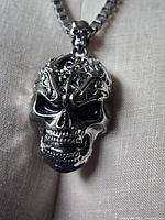 Close-up photo of a silver-tone skull pendant showing ornate detailed design on forehead and expressive facial features.