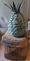 Full view showing decorative pineapple on top of wicker basket with geometric pattern.