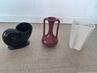 Three vases side by side on a flat surface, white background. Black shell-shaped spiral vase, burgundy double handled vase, white geometric vase.