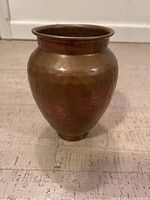 Front and side view of the hammered brass vase placed on a textured light-colored surface, showing its rounded shape, warm brass color and hammered texture.
