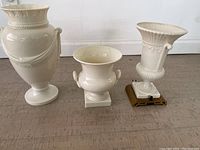 Three white ceramic Lenox items on the floor including two urns and one lamp base