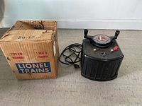 Black Lionel train transformer with dial, knob and attached cord, sitting on floor next to original box