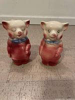 Front view of two ceramic Royal Copley piggy banks, pink with blue bow ties, one with closed eyes and the other with open eyes.