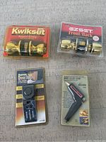 Four unopened miscellaneous household items displayed on a gray surface: two gold-tone Kwikset door knobs in packaging, one black super door stop alarm, and one Sears mini glue gun in original packaging.