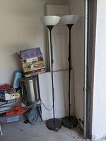 Two tall black metal floor lamps with white glass bowl shades standing next to a door in a cluttered space.