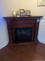 Full view of the wooden corner electric faux fireplace heater in a room corner, showing the cabinet and electric firebox insert with decorative items on top.