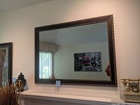 Front view of the large rectangular wall mirror hanging on a white wall above a mantle, showing dark wood frame with gold detailing and reflective glass surface.