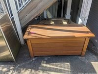 Front view of a rectangular outdoor wooden storage box with some shadows cast on it, placed outside near stairs and other outdoor items.
