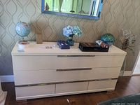 Front view of white lacquer sideboard with chrome handles, showing multiple drawers and decorative items on top including a globe, blue flowers, and ornamental glass orb.