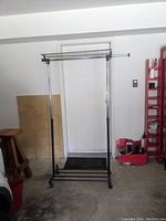 Full view of a vintage light duty clothes rack with a black metal frame, horizontal hanging bar, and caster wheels standing on a concrete floor in front of a white door.