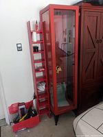 Red vintage light duty showcase cabinet with glass front and metal frame, showing overall design and included red narrow shelf with slots beside it. Cabinet stands on black legs and has gold-colored lock on door.