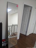 Full view of the tall rectangular mirror leaning against a wall near a doorway, showing its reflective surface and minimalist frame.