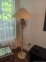 Full view of the vintage metal floor lamp standing near a window, showing the beige fabric lampshade and decorative base.