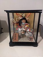 Front view of the Oriental doll in the glass display case, showing full figure, attire, headgear, and plaque.