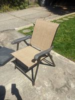 Photo of one folding lawn chair with tan mesh fabric and dark brown plastic armrests, metal frame, placed on outdoor pavement beside grass.