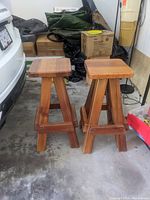 Pair of wooden stools with square tops placed on a concrete garage floor next to a car and storage items.