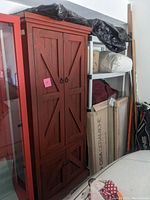Full view of the tall red rustic wooden cabinet standing beside shelving and other storage items.