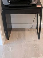Front view of small black desk with flat top surface and drawer closed. Positioned against wall with tile floor.