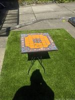 Full view of the small square table on artificial green grass, showcasing brightly colored tile top and black metal legs.