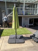 Full view of the closed green sun shade umbrella set on a paved outdoor patio with an artificial grass area in the background and residential home structure.