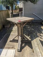 Tall wooden garden table outdoors on concrete patio, square plank top, sturdy cross-braced base, weathered wood surface