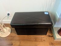 Full front view of black upholstered storage bench placed on wood floor next to an electrical outlet and floor lamp base.