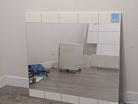Square wall mirror with white tiled frame standing vertically against a wall.