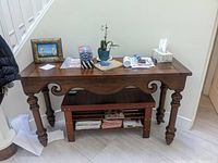 Full view of the wooden console sofa table placed against a wall under a staircase, with various papers and objects on top and a magazine rack underneath.
