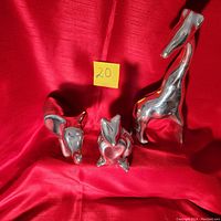 Front view of three aluminum animal sculptures on red cloth: elephant, kangaroo, giraffe. Noted size and shape differences. Shiny, polished metal with reflective surface.