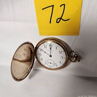 View of the open Waltham pocket watch showing the white dial with black numbers and a sub-second hand at 6 o'clock, gold-tone case with visible wear.