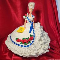 Front view of Barbie doll dated approx. 12 inches tall in white crochet dress with blue ribbon and red crocheted roses on a ruffled skirt.